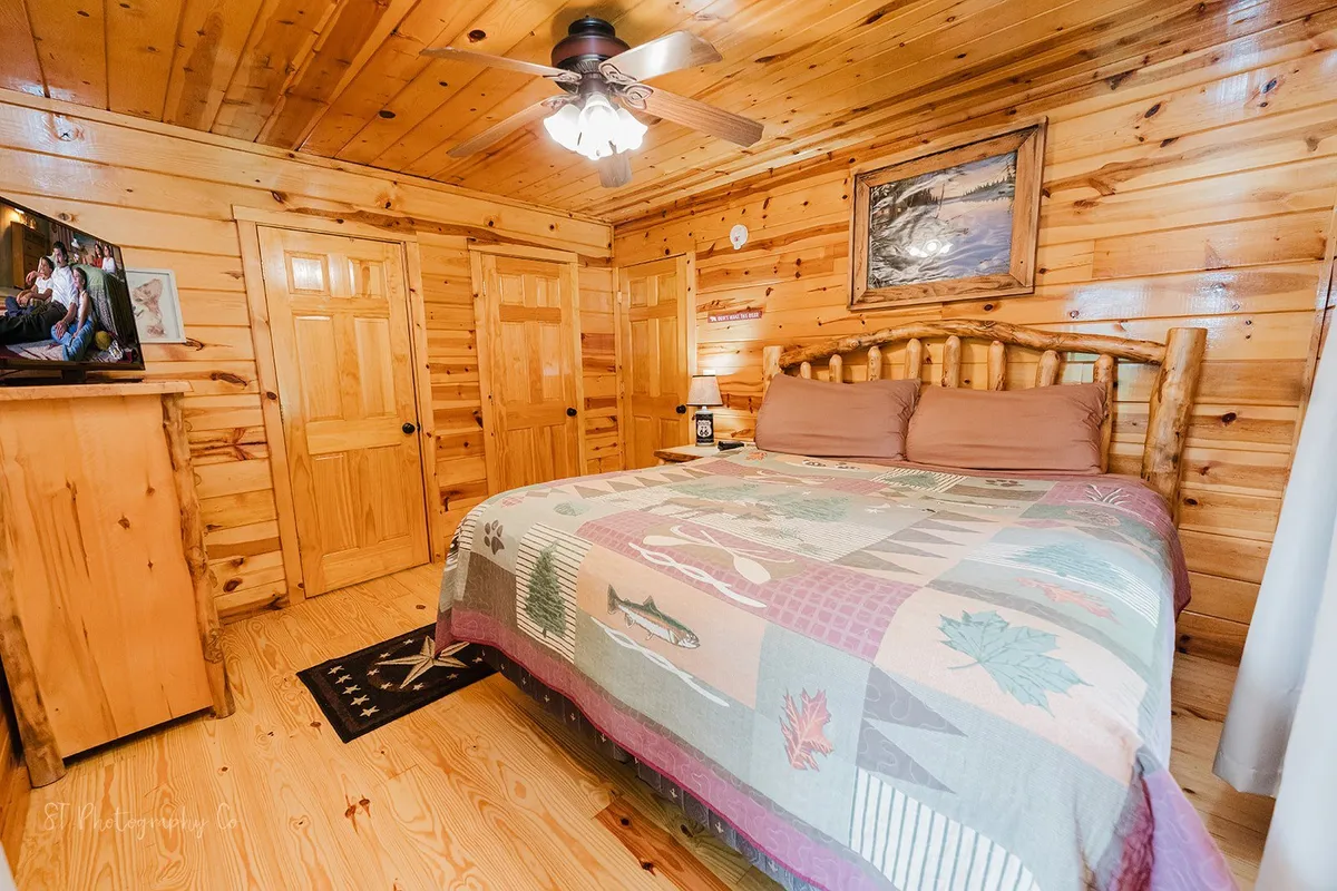 This image displays a cozy bedroom with a log bed, a colorful decorative quilt, wooden paneling, a television, and framed artwork, creating a welcoming and rustic ambiance.