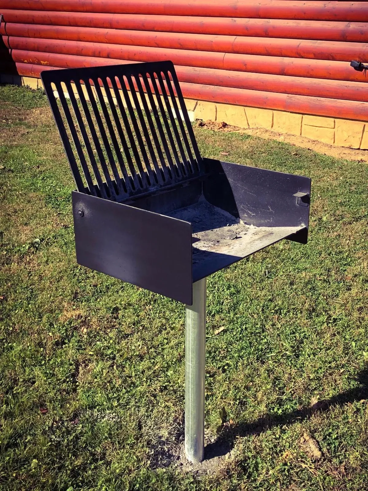A classic charcoal grill stands ready on a grassy patch beside the log cabin, with space for guests to gather and cook outdoors. The scene radiates a sense of relaxed, communal fun.