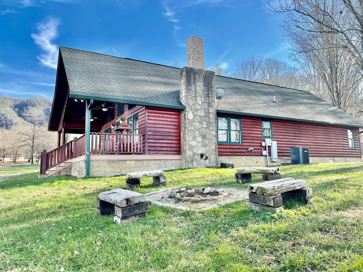 This image captures the log cabin’s exterior, focusing on the stone chimney and a fire pit area surrounded by seating. The porch is visible, set against a dramatic sunset sky, inviting guests to gather and unwind outside.
