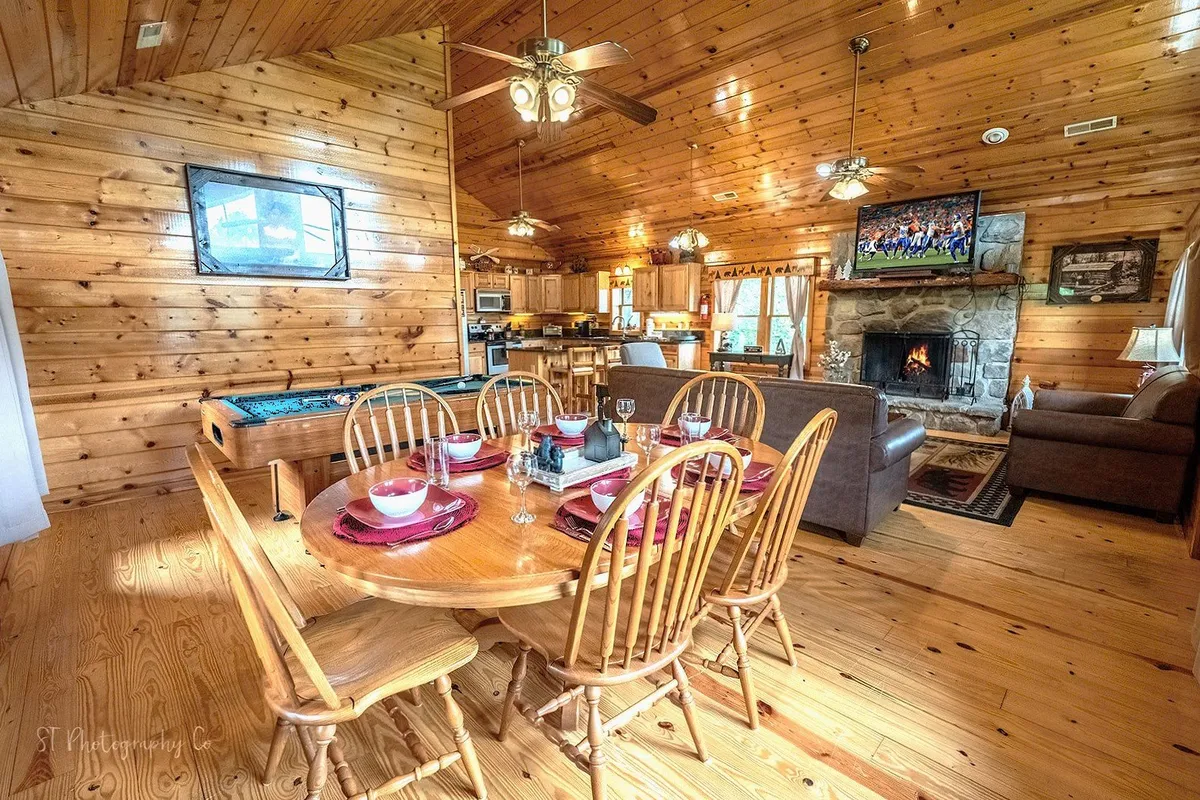 This scene showcases the living area’s connectivity, showing a dining table adjacent to the living space with a sofa, a fireplace, and a TV. The arrangement suggests versatility for both mealtimes and leisure activities like games or movies, all within a cozy, softly lit environment.