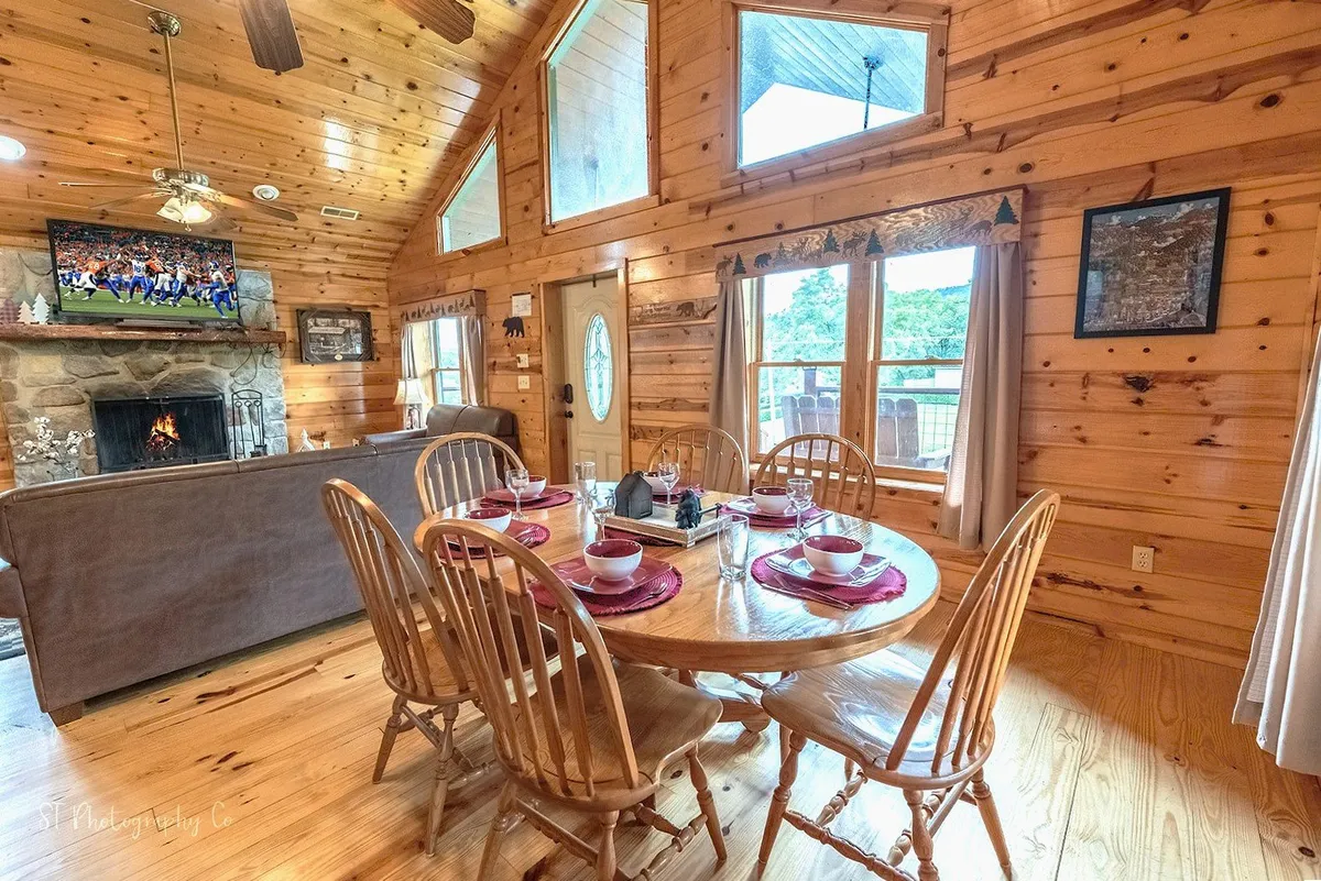 A dining table set for a meal sits in front of a roaring stone fireplace, with a television showing a sports game in the background. Wooden beams and rustic décor provide a cozy, communal setting.