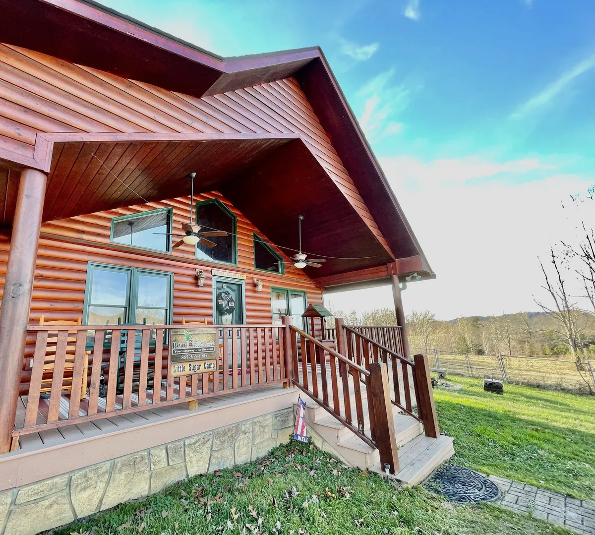 This view shows the log cabin’s charming exterior, with large windows, a welcoming wooden railing, and a signboard. The covered porch is in clear view, offering space for rocking chairs and morning relaxation.