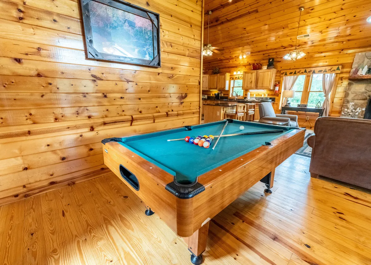 A casual and friendly entertainment area featuring a pool table, rustic wooden walls, decorative artwork, and a glimpse of the open kitchen. The space invites socializing and lively game nights, ideal for family entertainment.
