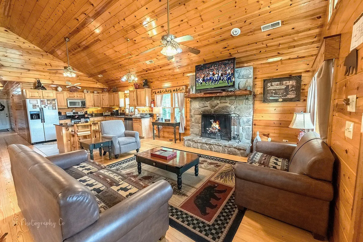 A different angle of the living room, this image features a comfortable sofa near the fireplace and a large flat-screen TV, with the adjoining kitchen visible in the background. The rustic touches and warm lighting enhance the space’s inviting atmosphere, blending modern amenities with cabin charm.