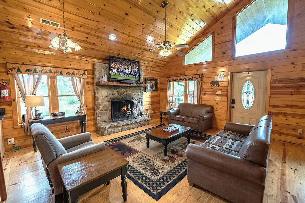This image presents a warm, inviting living room centered around a wood-burning fireplace. The plush sofa set and recliners are arranged for easy conversation and relaxation, with a television visible above the hearth and a cozy rug underfoot. The scene suggests a perfect spot for gathering, sharing stories, or enjoying a movie night.